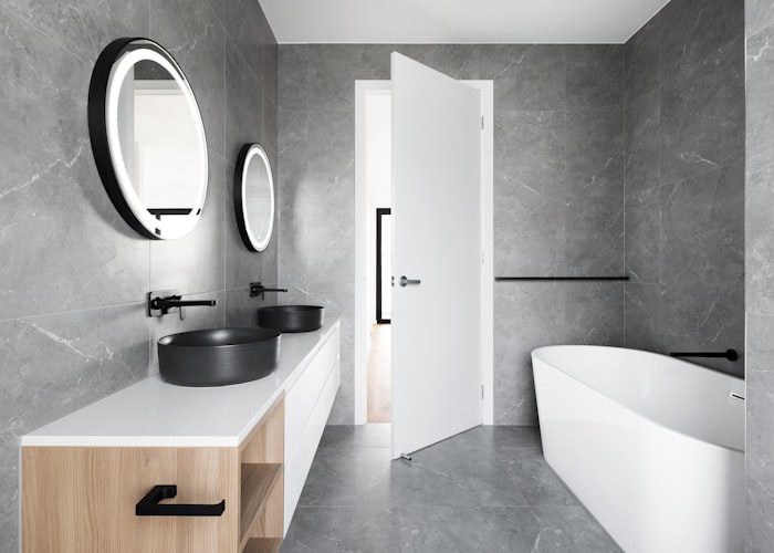Un baño moderno con paredes y suelo de microcemento gris, lavabo doble con espejos ovalados y bañera blanca.
