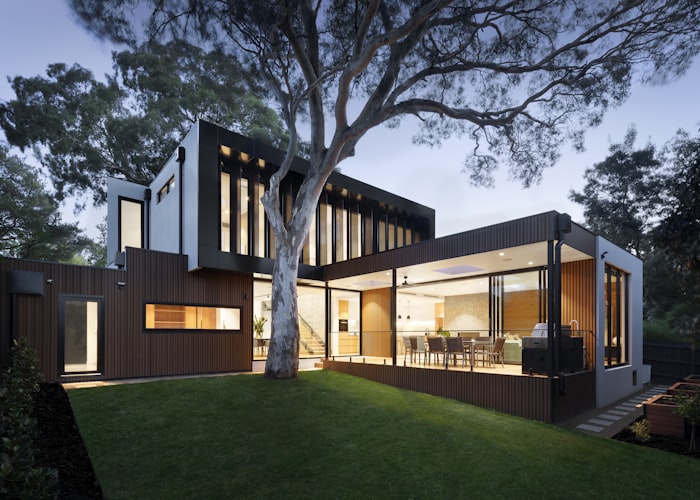 Una casa moderna con grandes ventanales y terraza iluminada al anochecer, rodeada de árboles y césped.
