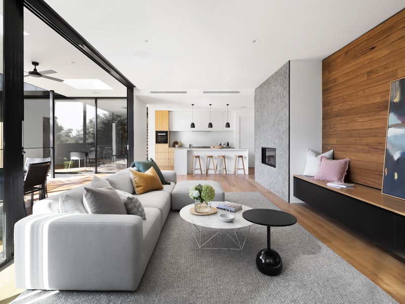 Salón moderno de planta abierta con sofá claro, cocina integrada y grandes ventanales al jardín.