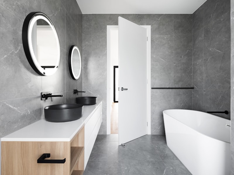 Baño moderno con paredes grises de acabado marmolado, lavabos negros, espejos ovalados y bañera exenta blanca.