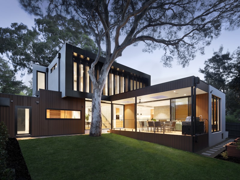 Una casa moderna acristalada aparece al atardecer, rodeada de árboles y con el interior iluminado con luz cálida.