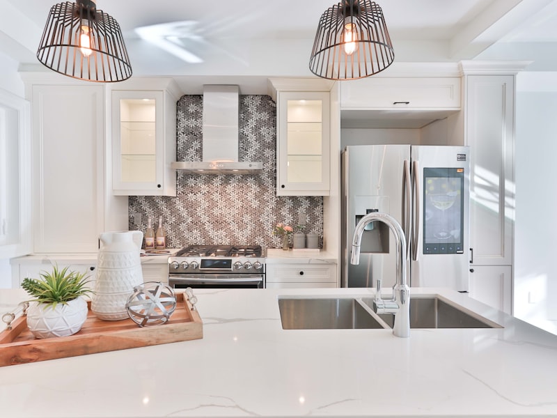 Una cocina blanca renovada en Sant Lluis con isla de cuarzo, nevera de acero y azulejos decorativos.