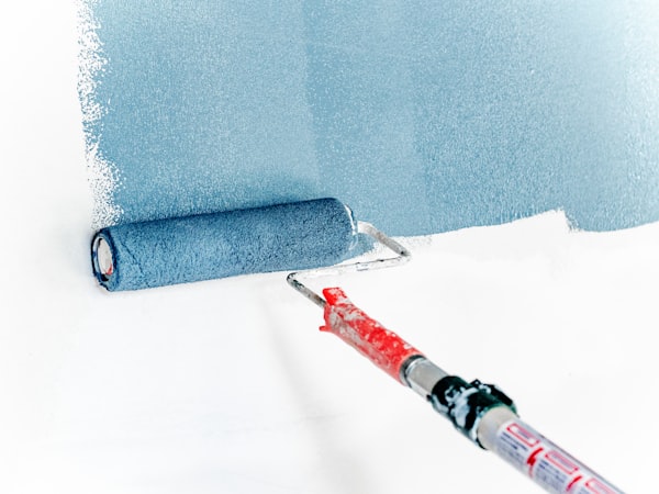 Un rodillo de pintura aplica una capa azul claro sobre una pared blanca, con el mango rojo visible.